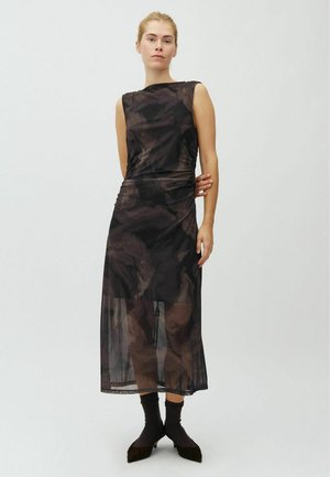 Frau steht in einem ärmellosen schwarzen gemusterten Midi-Kleid mit transparentem unterem Layer, trägt schwarze Socken und spitze schwarze Schuhe vor einfachem Hintergrund.