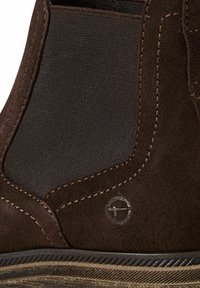 Brązowy zamszowy botek za kostkę z panelami tekstylnymi, kontrastowym przeszyciem oraz okrągłym logo w pobliżu podeszwy.