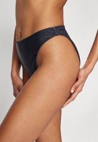 Aukšta juoda bikini kelnaitė su gūžta tekstūra, su lygiu juosmeniu ir aukštų išpjovų kojomis, sukurianti elegantišką siluetą.