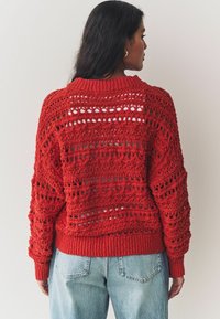 Pull en crochet rouge avec un design à mailles ouvertes et des poignets côtelés. Il présente un grand col et une coupe ample, associé à un jean bleu clair.