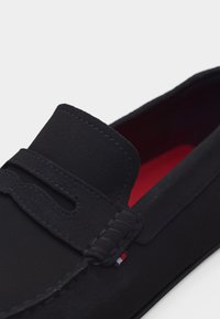 Μαύρο μοκασίνι από σουέτ με ραμμένες λεπτομέρειες, μύτη μοκασίνου και κόκκινη εσωτερική σόλα ορατή μέσα στο παπούτσι.