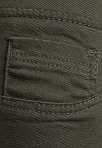 Mörk olivgrön denim med en liten bakficka. Har kontrasterande sömmar och en slät yta, typisk för material som används till avslappnade byxor.