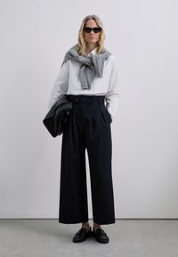 Hellebeine, dunkelblaue, gestreifte Hose mit hoher Taille und Doppelknopfdetails, kombiniert mit einem weißen Hemd und einem grauen Schal. Schwarze Slipper und Tasche.