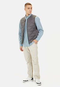Grijs gewatteerd vest over een lichtblauw overhemd met knopen, gecombineerd met beige broek en witte sneakers. Het vest heeft een ritssluiting en een golvenpatroon.