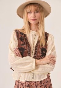 Mujer rubia con el cabello lacio, llevando un sombrero beige, blusa de encaje color crema, chaleco marrón bordado y falda de estampado, con los brazos cruzados.