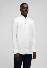 Witte overhemd met lange mouwen, gemaakt van textuurstof, met een button-down kraag en subtiele branding op de borst. Donkere spijkerbroek zichtbaar.