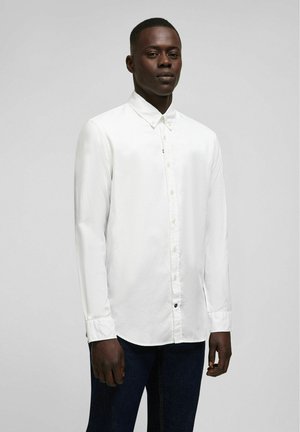 Weißes Langarmhemd aus strukturiertem Stoff, mit Button-Down-Kragen und dezenter Markenkennzeichnung auf der Brust. Dunkle Denim-Jeans sind sichtbar.