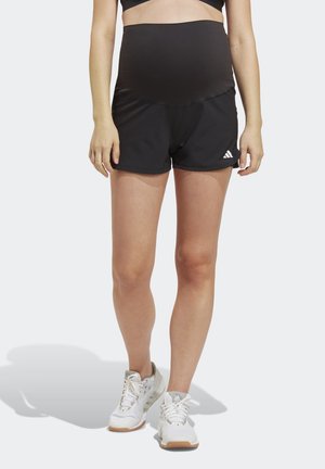 Femme portant un short de maternité noir avec le logo Adidas et des chaussures de sport blanches, debout devant un fond uni.