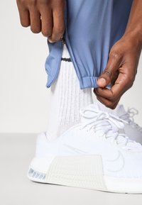 Μπλε αθλητικό παντελόνι με λεπτομέρεια φερμουάρ, λευκά υφασμάτινα sneakers και λευκές κάλτσες μέχρι τον αστράγαλο με μαύρη ρίγα κοντά στη μύτη.