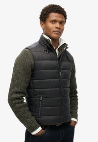 Schwarze Steppweste mit horizontalen Nähten, ausgestattet mit einem Reißverschluss vorne und seitlichen Taschen, über einem strukturierten grünen Pullover getragen.
