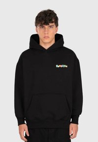 Jeune homme avec des cheveux bouclés portant un sweat à capuche noir avec un logo "dropsize" multicolore sur la poitrine, se tenant devant un fond gris clair.