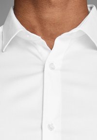 Camicia bianca con colletto strutturato, tessuto liscio e bottoni trasparenti. Presenta una vista ravvicinata del colletto e del décolleté.