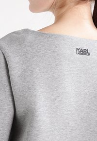 Sudadera de algodón gris con un escote amplio y un pequeño logo negro de "KARL LAGERFELD" en la parte superior de la espalda. Textura suave con un diseño casual.