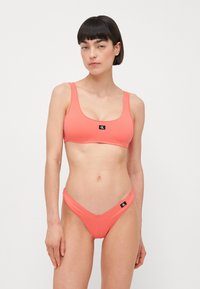 Korálový bikiny set s topom so štvorcovým strihom a širokými ramienkami, a zodpovedajúcimi nízkymi spodnými časťami. Každý kus má malú čiernu nálepku s logom.