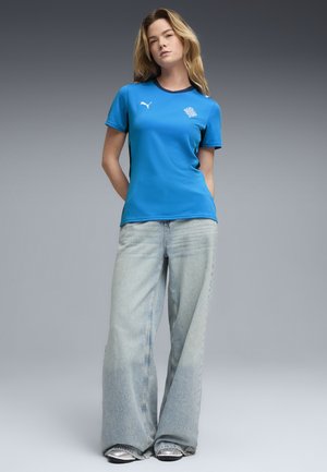 Camisa azul de manga corta con cuello redondo, que presenta un pequeño logo en el pecho, combinada con jeans de pierna ancha en azul claro y zapatos plateados.