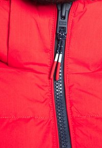 Rode pufferjack stof met een zwarte rits. Voorzien van dubbele trekbanden in rood en wit, en zichtbare stiksels op een gewatteerd oppervlak.
