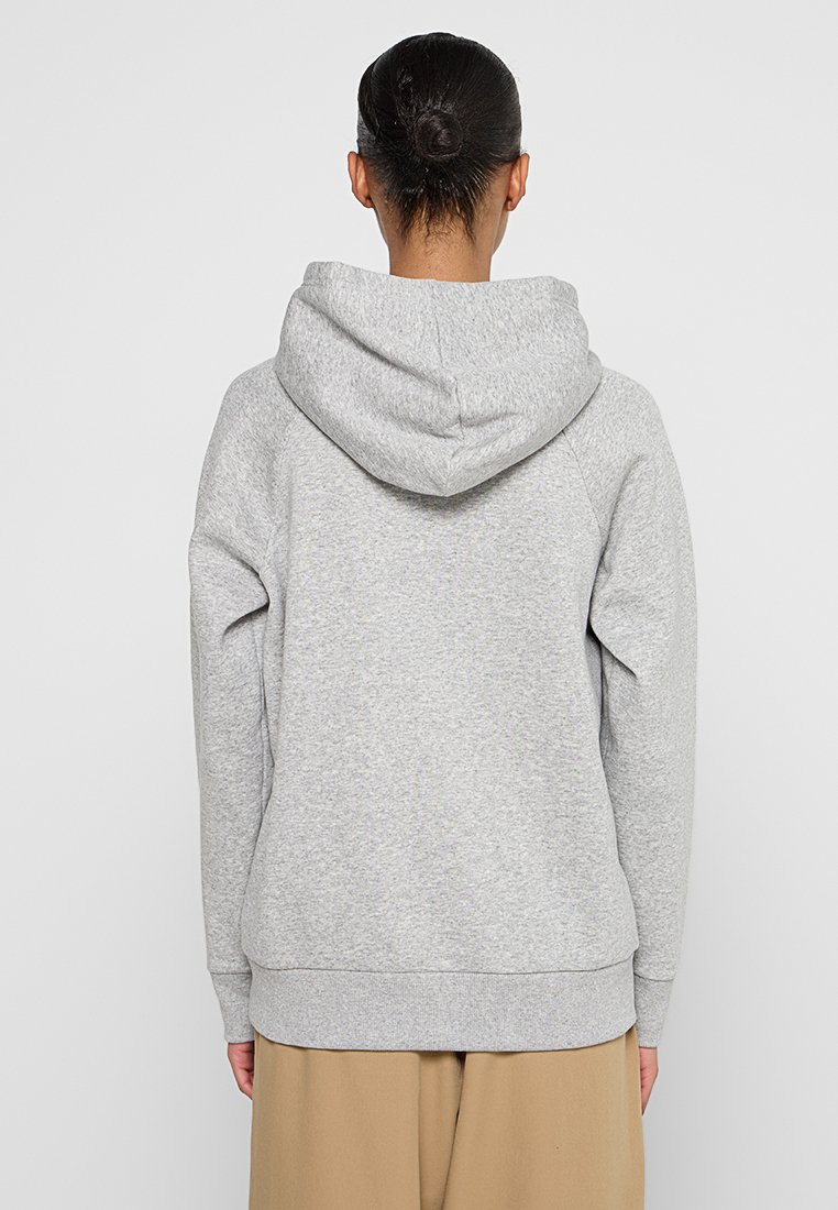 Mujer con el cabello oscuro recogido en un moño, vistiendo un sudadera con capucha gris claro y pantalones color beige, de pie de espaldas contra un fondo simple.