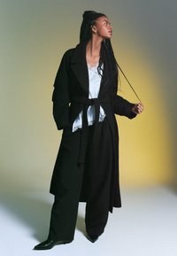 Svart bälte trenchcoat över en vit blus med spetsdetaljer, parat med vida byxor och svarta spetsiga klackar. Subtil textur och lager.