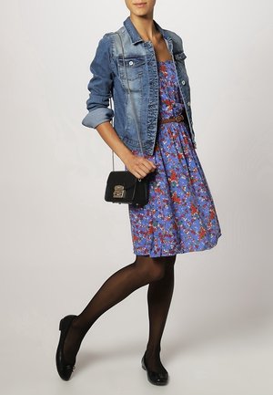 Jeansjacke über einem blauen Blumenkleid mit roten Akzenten, kombiniert mit schwarzen Strumpfhosen und Ballerinas sowie einer kleinen schwarzen Umhängetasche.