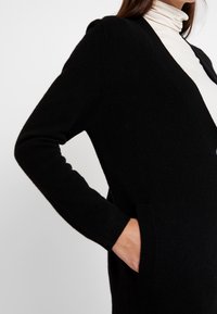 Svart stickad cardigan med långa ärmar, öppen framsida och sidofickor. Har en texturerad tyg och ribbade manschetter för extra detalj.