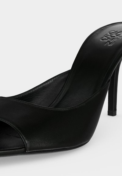 Μαύρο δερμάτινο mule με ψηλό τακούνι, ανοιχτό δάχτυλο και ανάγλυφο επώνυμο πάτο, εμφανίζεται σε απλό φωτεινό φόντο.