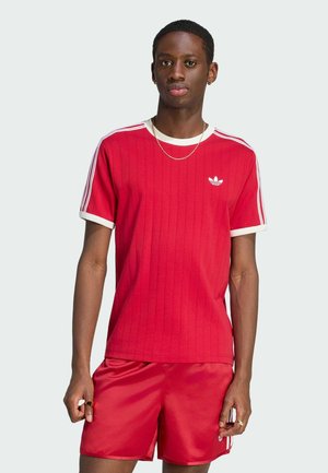 Young man wearing a red Adidas t-shirt with white stripes and matching red shorts, standing against a plain light background.