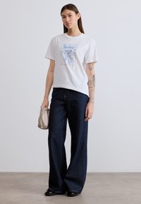 Λευκό βαμβακερό t-shirt με μπλε γραφικό τύπωμα, συνδυασμένο με σκούρες μπλε τζιν παντελόνες φαρδιάς γραμμής. Συμπληρώνεται με μικρή μπεζ τσάντα χειρός.