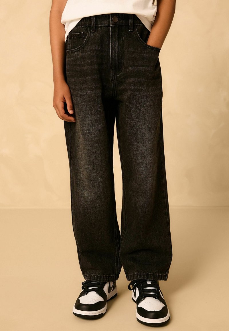 Person trägt lockere dunkle Jeans, schwarz-weiße Sneakers, mit einer Hand in der Tasche vor neutralem Hintergrund.