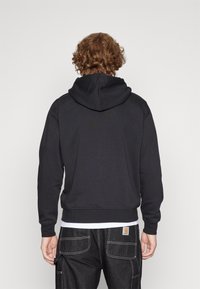 Uomo con capelli ricci che indossa un hoodie nero e pantaloni cargo neri con cuciture bianche, in piedi davanti a uno sfondo chiaro e semplice.