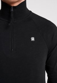 Svart zip-up långärmad skjorta med hög krage. Har en jämn textur, plana sömmar och en liten vit logotyp placerad på bröstet.