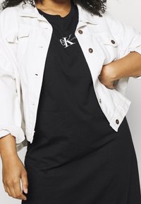 Biała dżinsowa kurtka z guzikami, założona na czarną koszulkę z logo. Częściowo widoczne kręcone włosy. Prosty, casualowy strój.