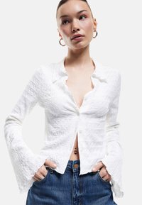 Weiße, strukturierte Bluse mit einem Kragen, Knopfverschluss und ausstellenden Ärmeln, kombiniert mit hochgeschnittenen Jeans.