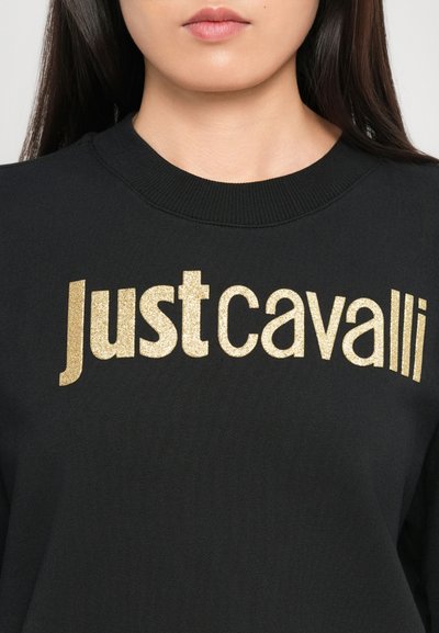 Femme portant un sweat-shirt noir à col rond avec un texte doré pailleté "Just Cavalli" à travers la poitrine.