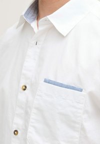 Witte katoenen shirt met een button-down kraag. Bevat een lichtblauwe accenten in de zak. Messing knopen, soepele textuur en een minimalistisch ontwerp.