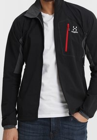 Svart softshell-jacka med grå accenter, ficka med dragkedja i brunt, hög krage och lätt design. Bärs över en vit t-shirt.