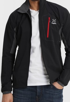 Zwarte softshell jas met grijze accenten, ritssluiting op de borstzak in het rood, hoge kraag en lichtgewicht ontwerp. Draag het over een wit T-shirt.