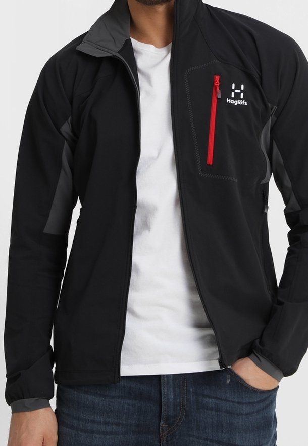 Svart softshell-jacka med grå accenter, ficka med dragkedja i brunt, hög krage och lätt design. Bärs över en vit t-shirt.