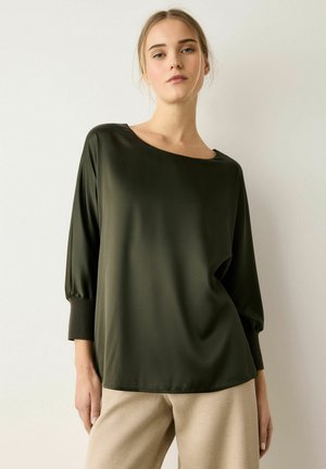 Junge Frau mit blonden Haaren trägt eine lockere dunkelgrüne Langarmbluse und eine hellbeige Hose und steht vor einem schlichten weißen Hintergrund.