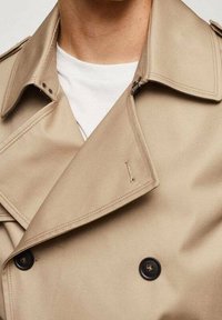 Beige trenchcoat met brede revers, dubbele knoopsluiting en zwarte knopen. Gladde stofstructuur met een subtiele glans.