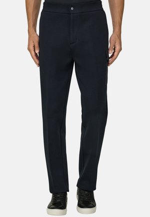 Uomo che indossa pantaloni neri a gamba dritta con tasca laterale e sneakers nere con suole bianche, in piedi davanti a uno sfondo semplice.