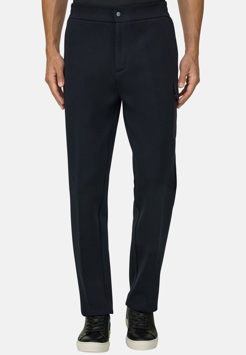 Homme portant un pantalon noir droit avec poche latérale et des baskets noires à semelles blanches, debout sur un fond uni.