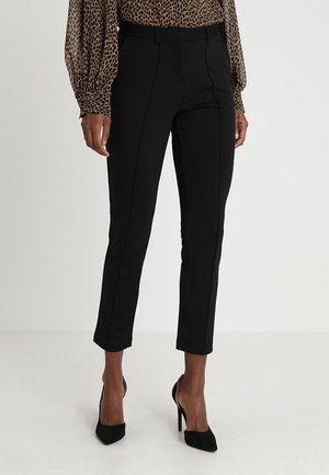 Femme portant un pantalon slim noir jusqu'à la cheville, un chemisier à manches longues imprimé léopard et des chaussures noires à talons aiguilles pointus, debout sur un sol blanc.