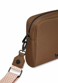 Braune Stoff-Umhängetasche mit strukturierter Oberfläche, schwarzem Reißverschluss und verstellbarem rosa Schulterriemen mit gewebtem Muster und Metallbeschlägen.