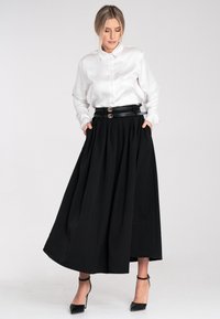 Weißes Satin-Hemd mit Knöpfen, kombiniert mit einem schwarzen plissierten Maxirock. Der Rock verfügt über einen schwarzen Gürtel mit doppelten goldenen Schnallen. Schwarze High Heels.