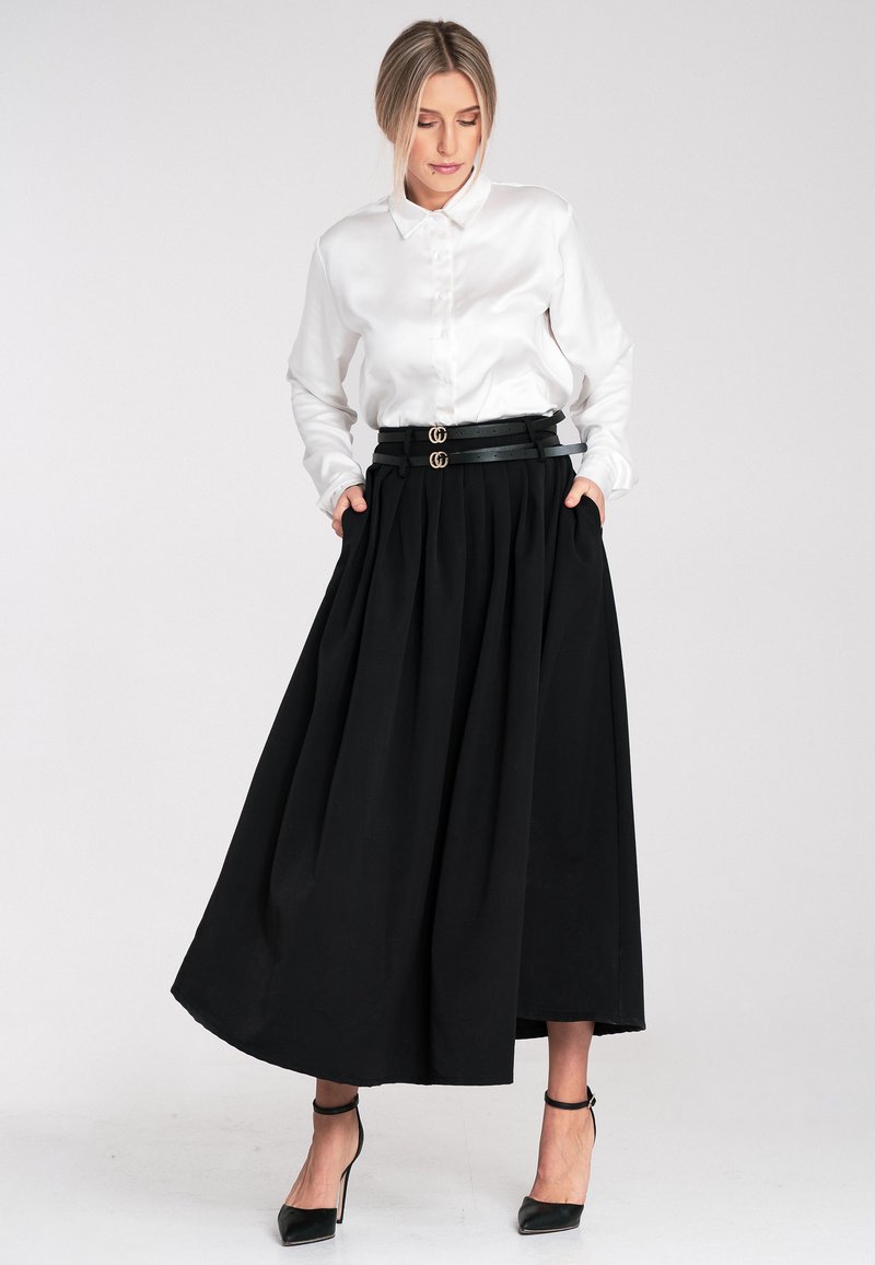 Weißes Satin-Hemd mit Knöpfen, kombiniert mit einem schwarzen plissierten Maxirock. Der Rock verfügt über einen schwarzen Gürtel mit doppelten goldenen Schnallen. Schwarze High Heels.