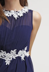 Námořnická modrá šaty s průsvitnou skládací látkou, s bílými květinovými krajkovými ozdobami kolem výstřihu a pasu pro přidaný detail.