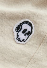 Un patch brodé représentant un crâne noir et blanc avec des écouteurs, sur un fond en tissu beige texturé.