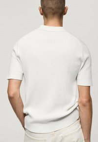 Polo blanc côtelé à manches courtes avec un col en maille plate et des manches retroussées, présentant une coupe ajustée et une texture subtile.