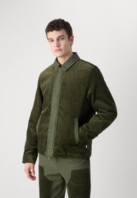 Green corduroy jacket with a collared neckline, snap buttons, and two side pockets. Features textured fabric and a relaxed fit design.