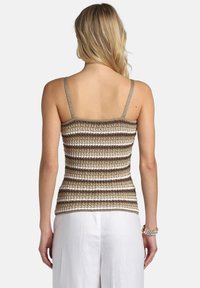 Mujer de cabello rubio vistiendo una blusa de tirantes de crochet a rayas marrón, blanca y beige, y pantalones blancos, vista desde atrás.